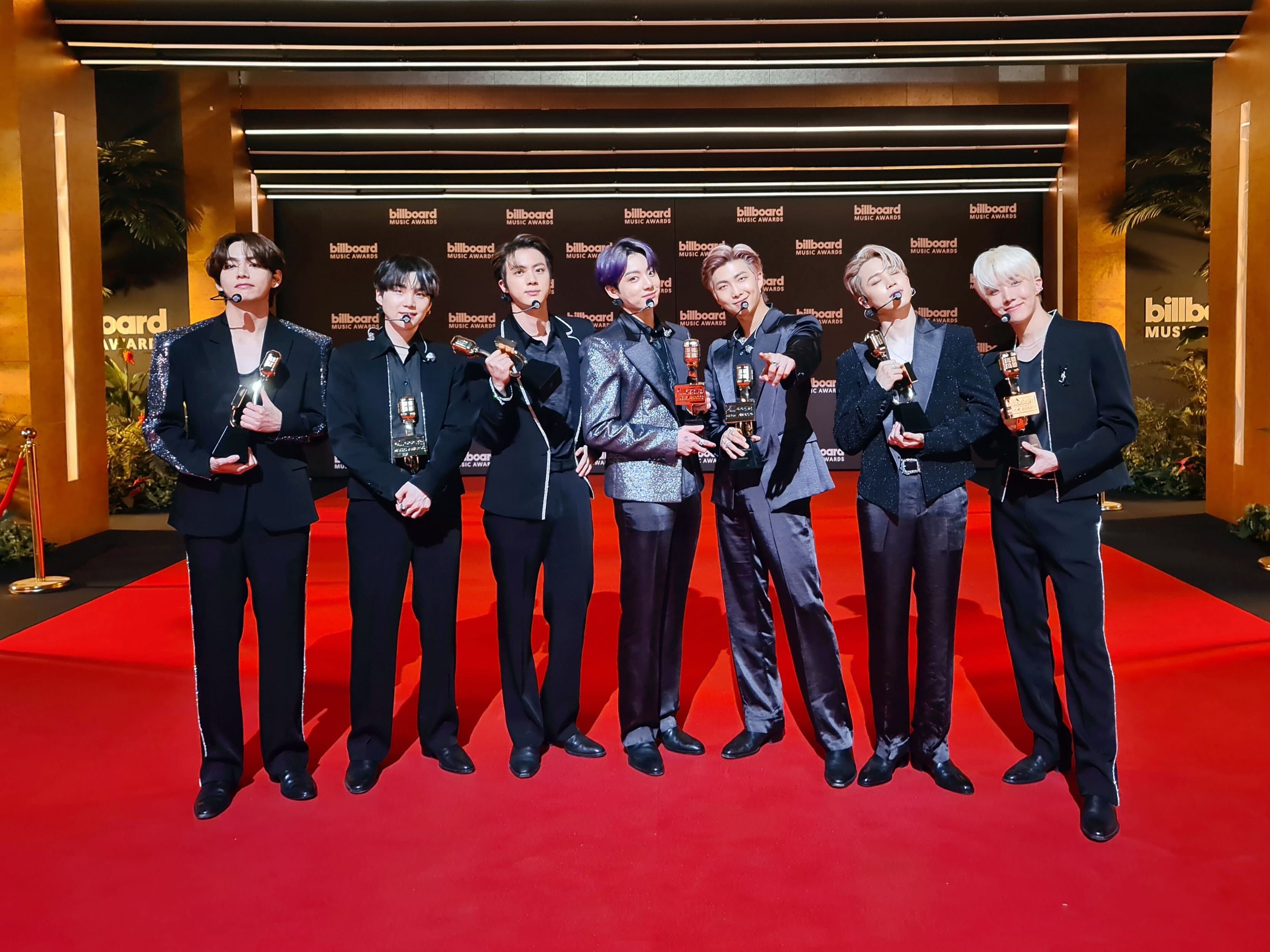 Seven members of BTS on the red carpet at the Billboard Music Awards 2021;  They are aligned horizontally, each holding a prize in a bold dress