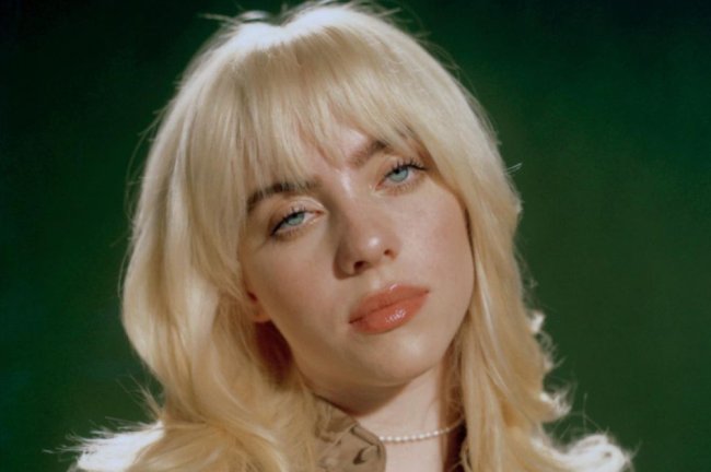 Billie Eilish posing seriously on a green background;  Her hair is down and she wears a beige shirt with a white pearl necklace.