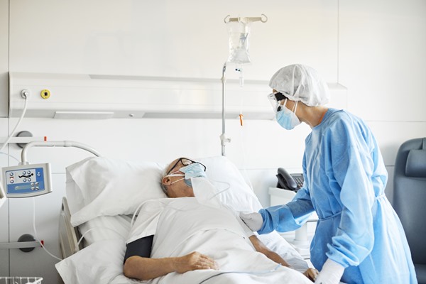 In the color photo, a person is lying on a hospital stretcher and another person is wearing blue with his hands on his arm.  Everyone wears masks