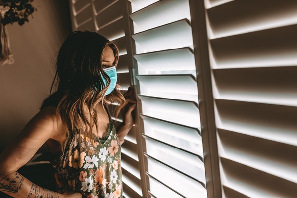 In the color photo, a woman looks out the window.  She is standing and wearing a mask