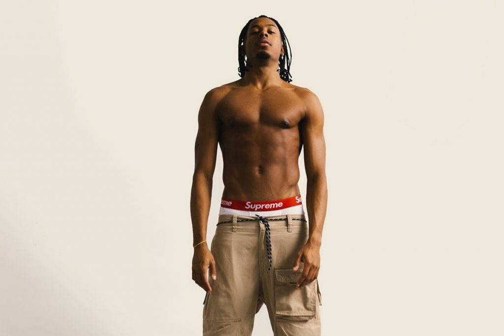 Former B.B. Paulo Andre, a tall black man with braided hair, poses for a photo on a white background.  He's shirtless and wears pants here with white underwear on display.  The underwear has a red stripe with the company logo, the top brand on it.