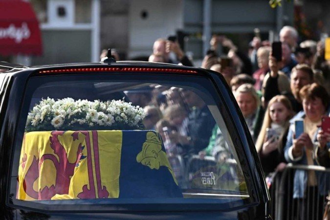 Members of the public pay their respects as they hearse carrying the coffin of Queen Elizabeth II, draped in the Royal Standard of Scotland, is driven through Ballater, on September 11, 2022. - Queen Elizabeth IIs coffin will travel by road through Scottish towns and villages on Sunday as it begins its final journey from her beloved Scottish retreat of Balmoral. The Queen, who died on September 8, will be taken to the Palace of Holyroodhouse before lying at rest in St Giles Cathedral, before travelling onwards to London for her funeral. (Photo by Paul ELLIS / AFP)        -  (crédito:  AFP)