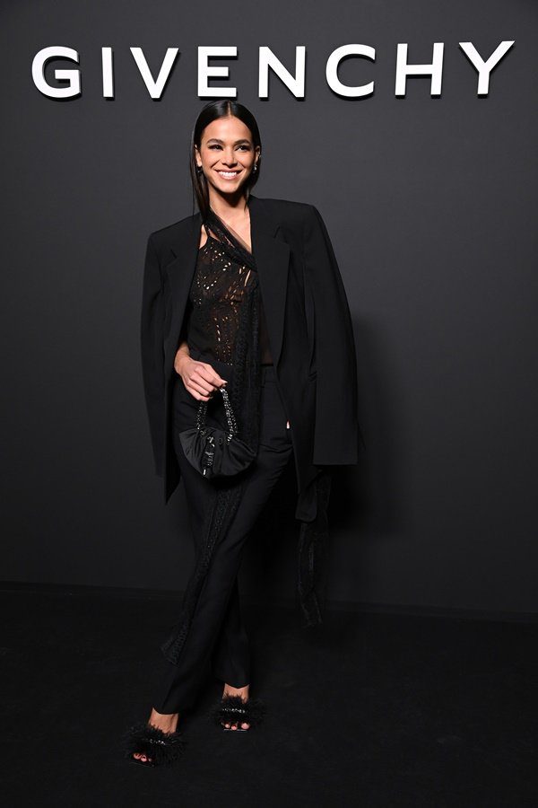 Actress Bruna Marquezine at the door of the Givenchy show at Paris Fashion Week.  She is a white young woman with long straight hair and wears a lace blouse, straight pants and a jacket.  All parts are black.